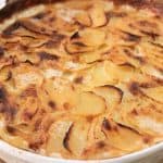 A round, white baking dish filled with a golden-brown potato gratin. The dish has a crispy, caramelized top layer of thinly sliced potatoes. The creamy texture and slight browning suggest it has been baked to a delicious finish. The dish rests on a wooden surface.