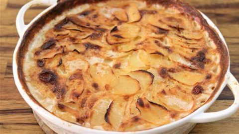 A round, white baking dish filled with a golden-brown potato gratin. The dish has a crispy, caramelized top layer of thinly sliced potatoes. The creamy texture and slight browning suggest it has been baked to a delicious finish. The dish rests on a wooden surface.