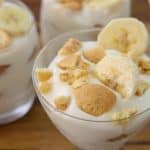 Four glasses of banana pudding are set on a wooden surface. Each glass is topped with banana slices and crumbled vanilla wafers, creating a layered dessert look. The creamy texture and golden brown pieces add appeal to this classic sweet treat.
