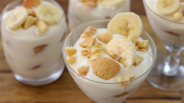 Four glasses of banana pudding are set on a wooden surface. Each glass is topped with banana slices and crumbled vanilla wafers, creating a layered dessert look. The creamy texture and golden brown pieces add appeal to this classic sweet treat.