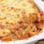 A close-up of a baked pasta dish with melted cheese on top in a white casserole dish. A portion has been scooped out, revealing creamy pasta with red sauce and visible chunks of tomato and herbs inside. The dish rests on a wooden surface.