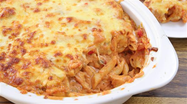 A close-up of a baked pasta dish with melted cheese on top in a white casserole dish. A portion has been scooped out, revealing creamy pasta with red sauce and visible chunks of tomato and herbs inside. The dish rests on a wooden surface.