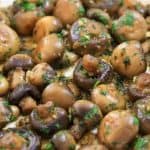 A close-up view of a dish featuring sautéed button mushrooms garnished with chopped parsley. The mushrooms are cooked to a golden brown and glisten with a buttery, herb-infused sauce. The vibrant green parsley contrasts with the rich, earthy tones of the mushrooms.