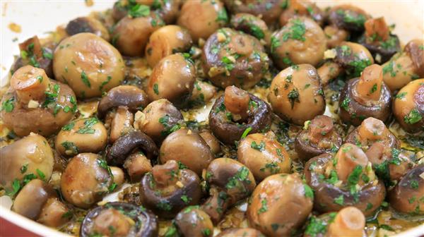 A close-up view of a dish featuring sautéed button mushrooms garnished with chopped parsley. The mushrooms are cooked to a golden brown and glisten with a buttery, herb-infused sauce. The vibrant green parsley contrasts with the rich, earthy tones of the mushrooms.