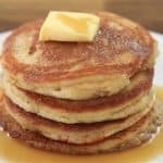 A stack of four golden-brown pancakes sits on a white plate, topped with a square of melting butter. Syrup has been drizzled over the pancakes, pooling around them on the plate. The background shows a wooden table surface.