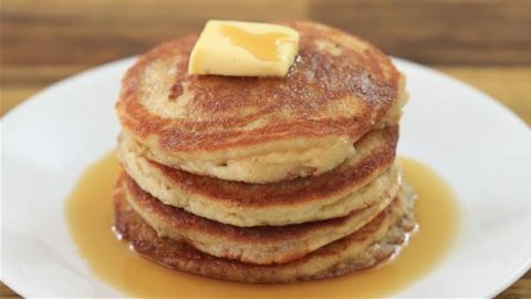 A stack of four golden-brown pancakes sits on a white plate, topped with a square of melting butter. Syrup has been drizzled over the pancakes, pooling around them on the plate. The background shows a wooden table surface.