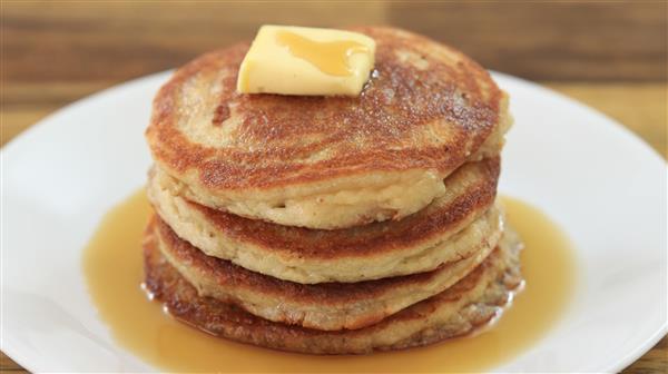 A stack of four golden-brown pancakes sits on a white plate, topped with a square of melting butter. Syrup has been drizzled over the pancakes, pooling around them on the plate. The background shows a wooden table surface.