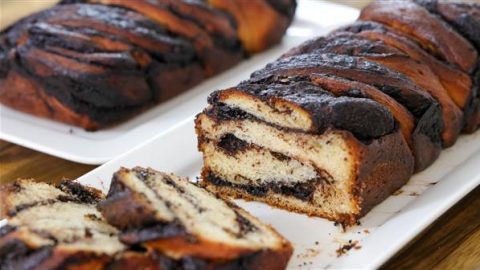 A loaf of chocolate babka is sliced on a rectangular white plate, revealing its swirled chocolate filling. Another intact loaf sits in the background on a similar plate. The golden-brown crust contrasts with the rich, dark chocolate interior.