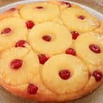 A round pineapple upside-down cake sits on a clear glass plate. The cake is adorned with rings of pineapple and maraschino cherries, each cherry placed in the center of the pineapple rings and in between them, giving it a vibrant and inviting appearance.