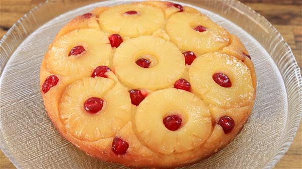 A round pineapple upside-down cake sits on a clear glass plate. The cake is adorned with rings of pineapple and maraschino cherries, each cherry placed in the center of the pineapple rings and in between them, giving it a vibrant and inviting appearance.