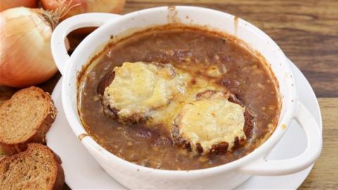 A bowl of French onion soup topped with melted cheese-covered bread slices. The soup is served in a white ceramic dish, placed on a wooden surface with sliced baguette pieces and whole onions in the background.