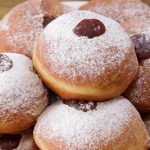 A plate piled with jelly-filled donuts dusted with powdered sugar. The golden brown donuts have visible dollops of red jelly peeking out from the centers, creating a delectable and inviting presentation. The background features a wooden surface.