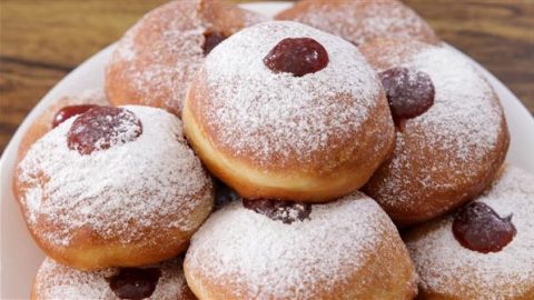 A plate piled with jelly-filled donuts dusted with powdered sugar. The golden brown donuts have visible dollops of red jelly peeking out from the centers, creating a delectable and inviting presentation. The background features a wooden surface.