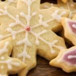 Assorted snowflake-shaped cookies decorated with white and pink icing are arranged on a wooden surface. One cookie features a small pink pearl at its center.