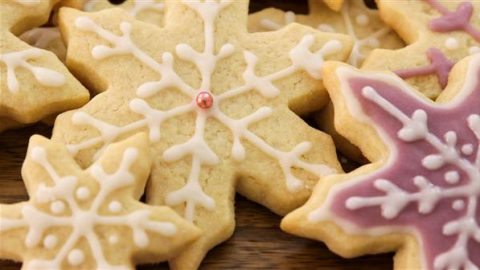 Assorted snowflake-shaped cookies decorated with white and pink icing are arranged on a wooden surface. One cookie features a small pink pearl at its center.