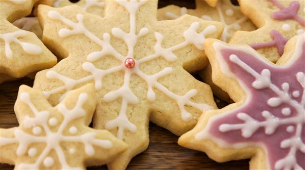 Assorted snowflake-shaped cookies decorated with white and pink icing are arranged on a wooden surface. One cookie features a small pink pearl at its center.