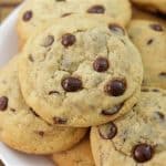 A close-up of a plate of chocolate chip cookies piled on top of each other. The cookies appear golden-brown and have visible chocolate chips. The plate is against a wooden background.