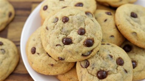 A close-up of a plate of chocolate chip cookies piled on top of each other. The cookies appear golden-brown and have visible chocolate chips. The plate is against a wooden background.