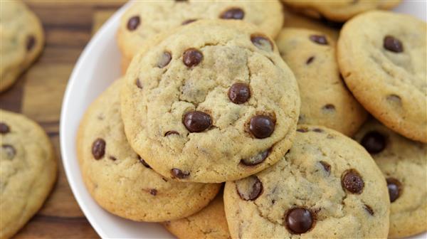 A close-up of a plate of chocolate chip cookies piled on top of each other. The cookies appear golden-brown and have visible chocolate chips. The plate is against a wooden background.