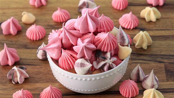 A small white bowl is filled with an assortment of colorful meringue kisses, predominantly in shades of pink, purple, and white. More meringues are scattered around the wooden surface, adding to the festive and sweet presentation.