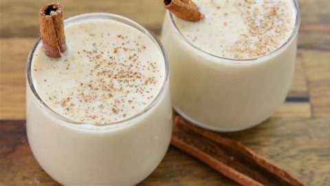 Two glasses filled with creamy eggnog are topped with ground nutmeg and a cinnamon stick. The drinks are set on a wooden surface with two additional cinnamon sticks lying beside the glasses, evoking a cozy holiday feel.