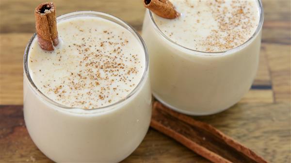 Two glasses filled with creamy eggnog are topped with ground nutmeg and a cinnamon stick. The drinks are set on a wooden surface with two additional cinnamon sticks lying beside the glasses, evoking a cozy holiday feel.