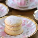 A decorative plate with three powdered sugar-coated cookies is placed on a wooden table. In the background are additional plates with cookies and a teacup filled with tea, all featuring a matching floral and gold design.