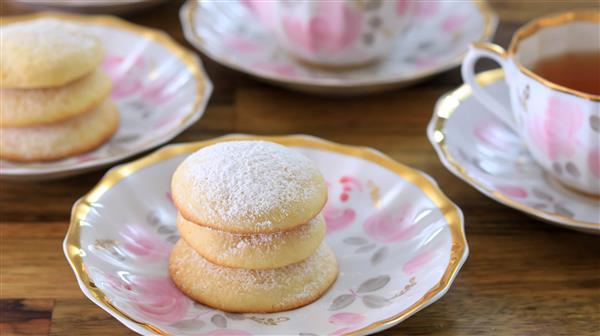 A decorative plate with three powdered sugar-coated cookies is placed on a wooden table. In the background are additional plates with cookies and a teacup filled with tea, all featuring a matching floral and gold design.