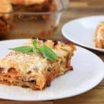 A serving of lasagna topped with grated cheese and basil sits on a white plate. Another piece of lasagna on a similar plate is in the background, near a glass baking dish with the remaining lasagna. The scene is set on a wooden surface.