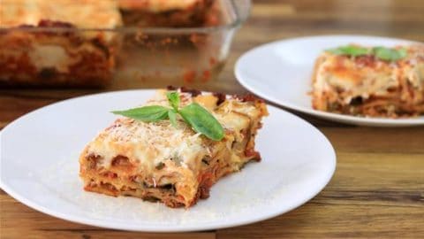 A serving of lasagna topped with grated cheese and basil sits on a white plate. Another piece of lasagna on a similar plate is in the background, near a glass baking dish with the remaining lasagna. The scene is set on a wooden surface.