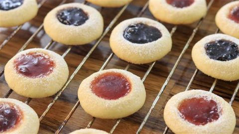 A cooling rack holds several jam-filled thumbprint cookies. The cookies have a light dusting of powdered sugar and are filled with different jams, with some centers appearing red and others a darker purple. The wooden surface underneath adds a rustic feel.