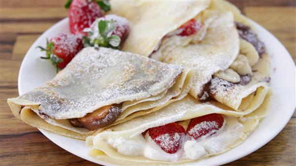 A plate of filled crepes dusted with powdered sugar. The crepes are folded and contain various fillings, including fresh strawberries, whipped cream, and possibly chocolate or banana slices. Some whole strawberries are also placed on the plate for garnish.