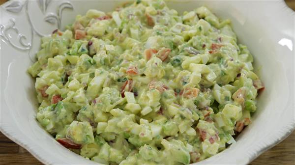 A close-up of a bowl filled with creamy avocado egg salad. The dish contains chopped avocado, hard-boiled eggs, tomatoes, and a creamy dressing, all mixed together. The white bowl has a decorative pattern on the rim.