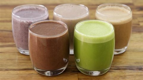 Five glasses of smoothies are arranged in two rows on a wooden surface. The glasses contain different colored smoothies: two chocolate brown, one green, one purple, and one beige.