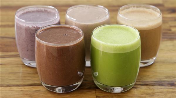 Five glasses of smoothies are arranged in two rows on a wooden surface. The glasses contain different colored smoothies: two chocolate brown, one green, one purple, and one beige.