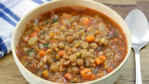 A bowl of hearty lentil soup filled with lentils, diced carrots, onions, and herbs, placed on a wooden surface. A silver spoon is positioned to the right, and a blue and white striped cloth napkin is partially visible on the left.