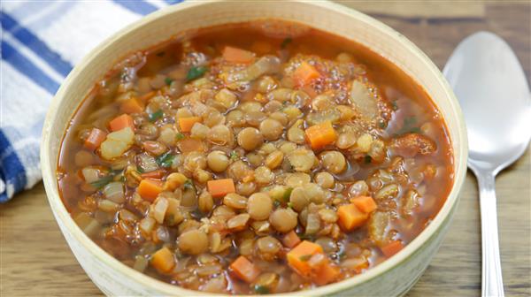 A bowl of hearty lentil soup filled with lentils, diced carrots, onions, and herbs, placed on a wooden surface. A silver spoon is positioned to the right, and a blue and white striped cloth napkin is partially visible on the left.