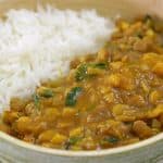 A bowl filled with white rice and a serving of lentil curry. The curry is yellow-orange in color and includes visible chunks of vegetables and green herbs. The bowl is placed on a wooden surface.