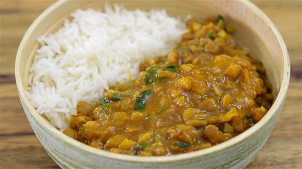 A bowl filled with white rice and a serving of lentil curry. The curry is yellow-orange in color and includes visible chunks of vegetables and green herbs. The bowl is placed on a wooden surface.