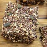 Close-up shot of a stack of homemade multi-seed crackers on a wooden surface. The crackers are packed with a variety of seeds, including flax, sesame, pumpkin, and sunflower seeds, giving them a dense, crunchy texture.