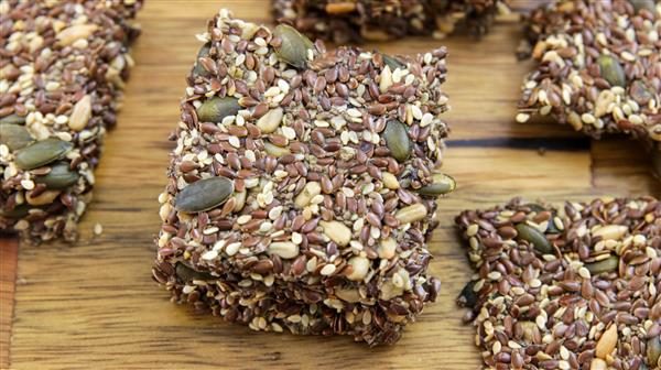 Close-up shot of a stack of homemade multi-seed crackers on a wooden surface. The crackers are packed with a variety of seeds, including flax, sesame, pumpkin, and sunflower seeds, giving them a dense, crunchy texture.