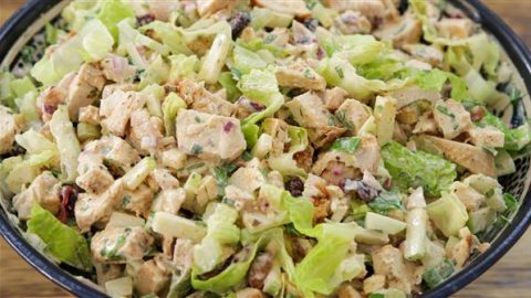 A close-up of a bowl filled with a hearty chicken salad. It contains diced grilled chicken, chopped lettuce, red onions, celery, raisins, and is dressed with a creamy sauce. The vibrant colors and diverse textures of the ingredients are clearly visible.