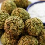 A plate of golden-brown falafel balls is arranged around a small blue dish filled with creamy white dipping sauce. The falafel is crispy on the outside, indicating it is freshly fried, with a texture that showcases the mixture of herbs and chickpeas.