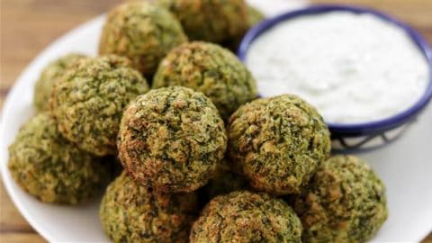 A plate of golden-brown falafel balls is arranged around a small blue dish filled with creamy white dipping sauce. The falafel is crispy on the outside, indicating it is freshly fried, with a texture that showcases the mixture of herbs and chickpeas.