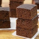 Three neatly stacked pieces of chocolate brownies on a wooden surface with more brownies in the background. Each square piece has a rich, dense texture and dark chocolate color, placed on small white parchment papers.