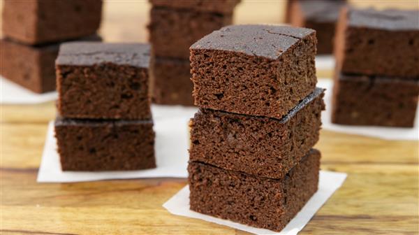 Three neatly stacked pieces of chocolate brownies on a wooden surface with more brownies in the background. Each square piece has a rich, dense texture and dark chocolate color, placed on small white parchment papers.