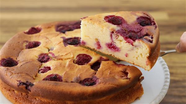 A close-up of a cheesecake with a golden-brown crust and topped with what appears to be cherries or berries. A hand is lifting a slice from the cake, revealing a creamy texture with berry pieces inside. The cake rests on a white plate, and the background is blurred.