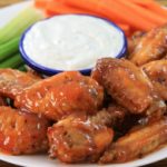 A plate of glazed chicken wings served with a side of celery and carrot sticks. A small blue-rimmed bowl filled with white dipping sauce is placed in the center of the plate. The dish is set on a wooden surface.