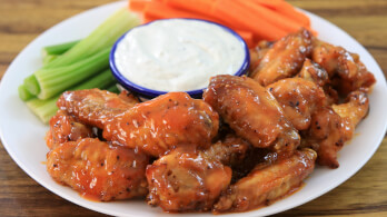 A plate of glazed chicken wings served with a side of celery and carrot sticks. A small blue-rimmed bowl filled with white dipping sauce is placed in the center of the plate. The dish is set on a wooden surface.