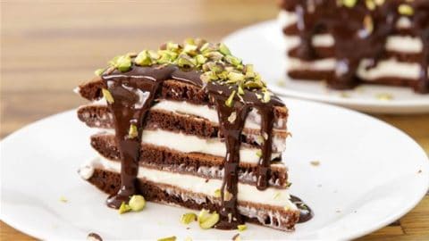 A slice of chocolate layer cake with white cream filling between the layers, topped with rich chocolate sauce and garnished with chopped pistachios, served on a white plate. Another slice is visible in the background on a separate plate.